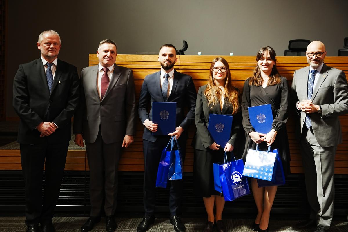 Grupa czterech mężczyzn i dwóch kobiet pozuje do zdjęcia przed drewnianym biurkiem. Jeden mężczyzna i dwie kobiety trzymają w dłoniach niebieskie okładki z wizerunkiem złotego orła oraz dwie niebieskie torebki prezentowe.