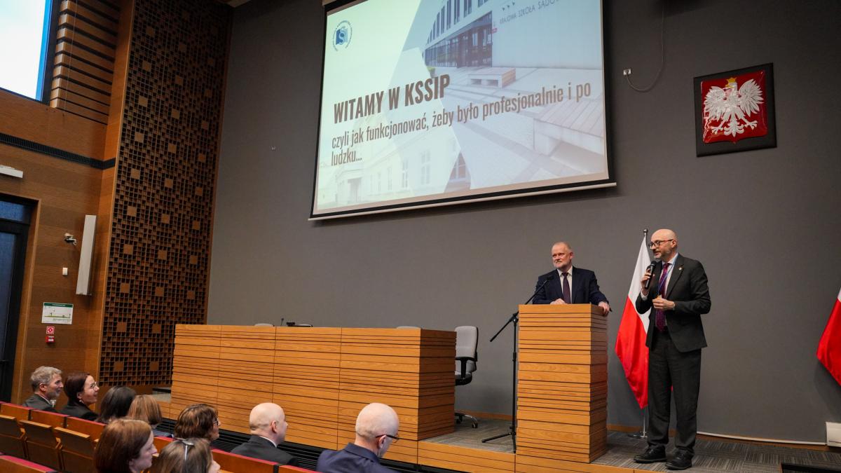 Na zdjęciu na podwyższeniu przy mównicy stoi dwóch mężczyzn, z twarzami zwróconymi w stronę audytorium. Jeden z nich trzyma w ręku mikrofon, zwracając się w stronę widowni, na której widać siedzące osoby. Za plecami stojących mężczyzn widać ekran powitalny, godło państwa oraz dwie flagi Polski. Na ekranie widoczny jest slajd z napisem „Witamy w KSSiP, czyli jak funkcjonować , żeby było profesjonalnie i po ludzku”.