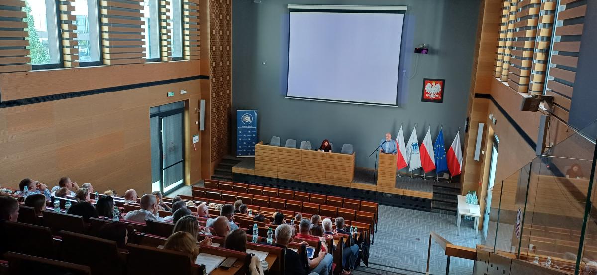 Grupa około sześćdziesięciu osób: kobiet i mężczyzn siedzi na dużej auli wykładowej. Przed nimi, na podeście, przy dużym biurku siedzi kobieta. Obok, przy mównicy stoi mężczyzna. W tle po prawej stronie widać godło Polski oraz flagi Polski, Unii Europejskiej i Krajowej Szkoły Sądownictwa i Prokuratury. Po lewej stronie znajduje się roll-up z logo Krajowej Szkoły Sądownictwa i Prokuratury.