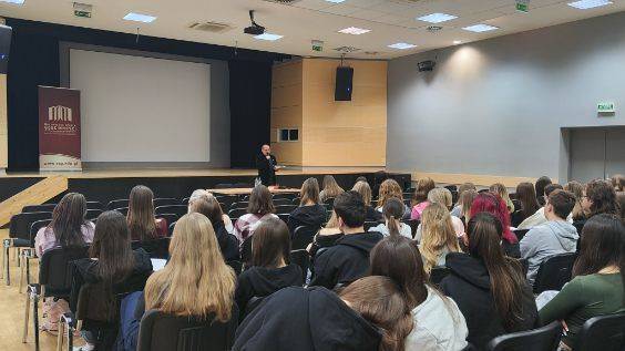 grupa ludzi siedzących. Przed nimi stoi mężczyzna z mikrofonem. W tle rzutnik oraz roll up z logiem Zespołu Szkół - Małopolskiej Szkoły Gościnności